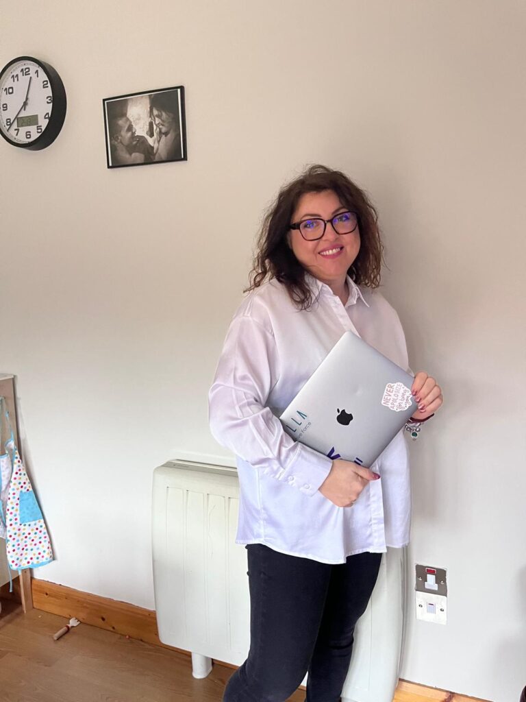 Kiki Paschalidou holding a laptop with Aella Creative Force branding in a bright workspace.