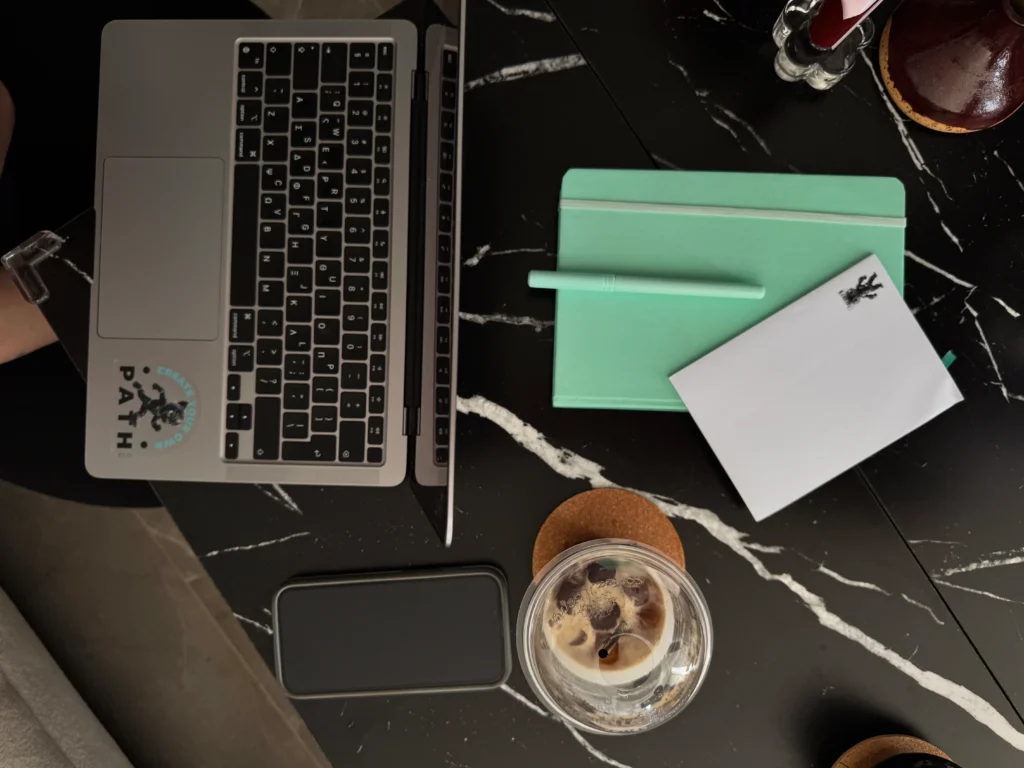 Overhead flat lay of a creative workspace on a black marble surface with an open MacBook laptop, mint green notebook, white notepad, phone, and iced coffee on a cork coaster